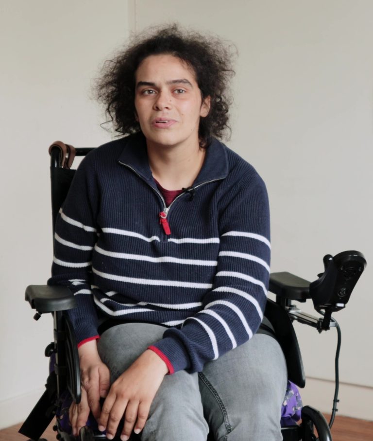 Photo of Ashleigh, featured in the video for Disability Awareness Sunday 2023. She has black curly hair, and is sat in her wheelchair, wearing a blue and white stripey jumper.